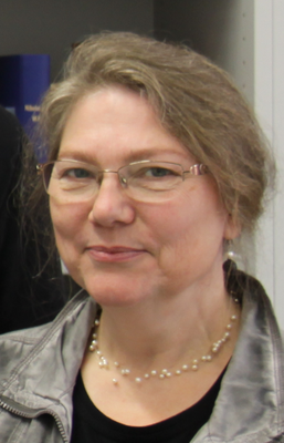 eine Frau mit Brille, halblangem braungrauem Haar und Halskette blickt verschmitzt in die Kamera